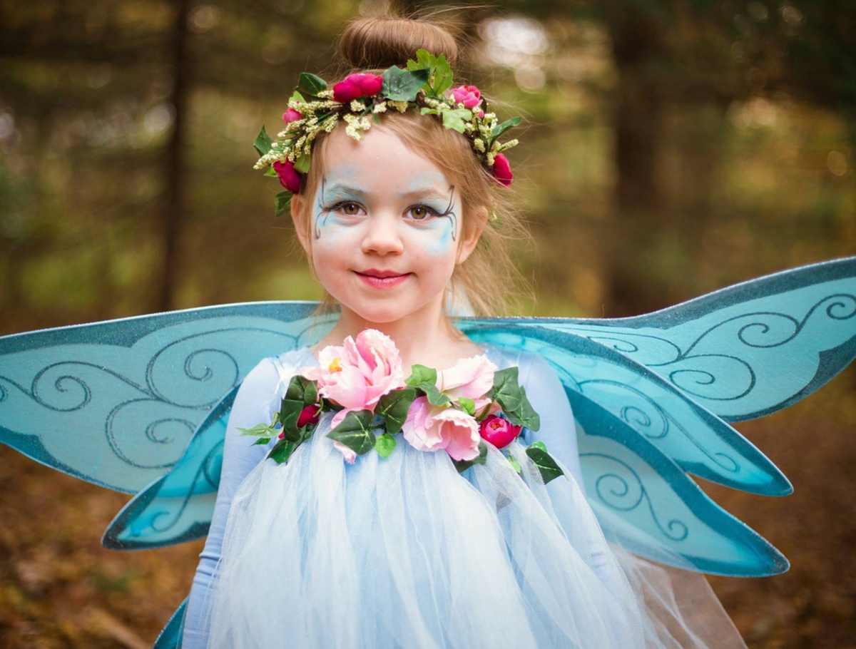 A young girl dressed as a fairy stands outdoors in a forest, wearing a light blue tulle costume with translucent blue wings, pink flowers, green leaves, and a floral crown.
